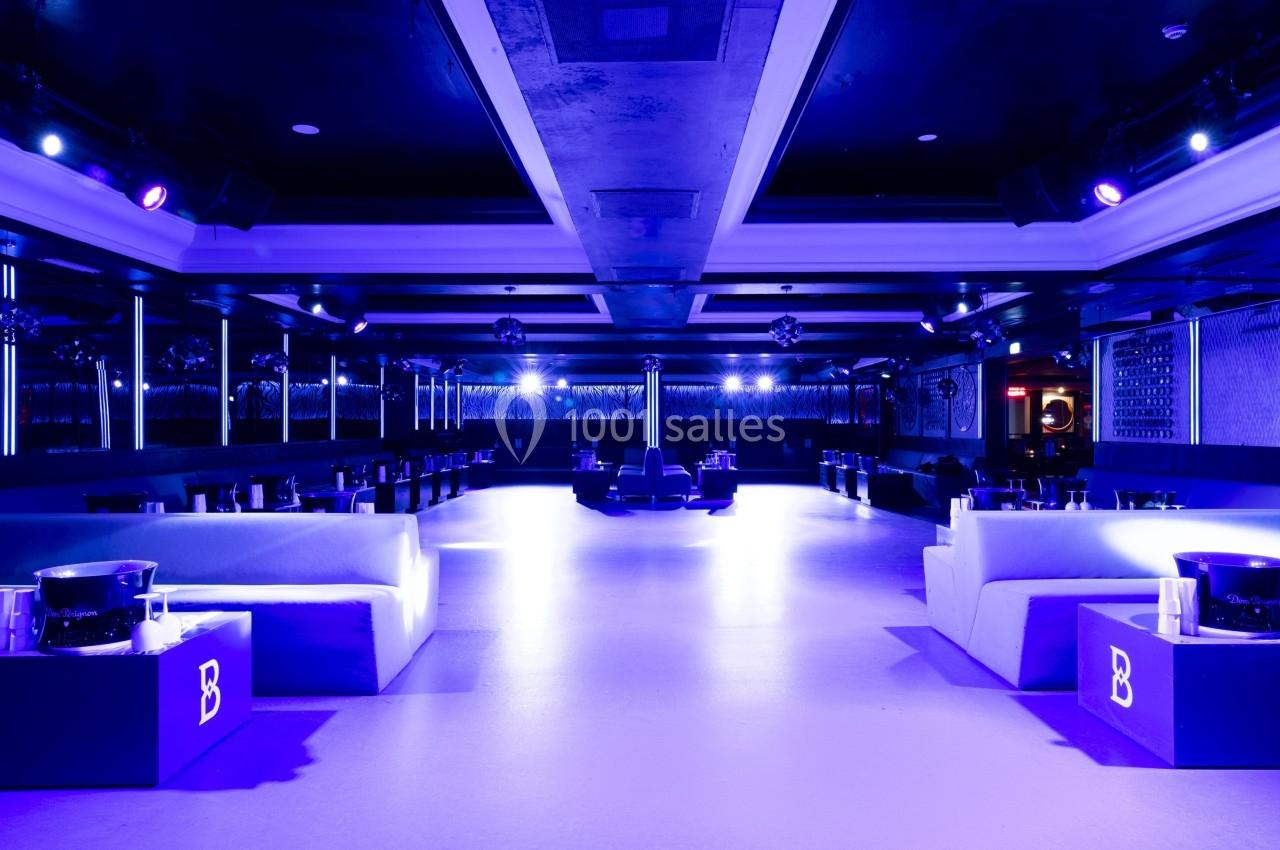 Salle de réception moderne avec éclairage violet, banquettes alignées et tables basses sur un sol clair.