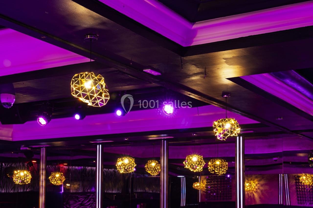 Plafond d'une salle avec des luminaires géométriques dorés et un éclairage violet ambiant.