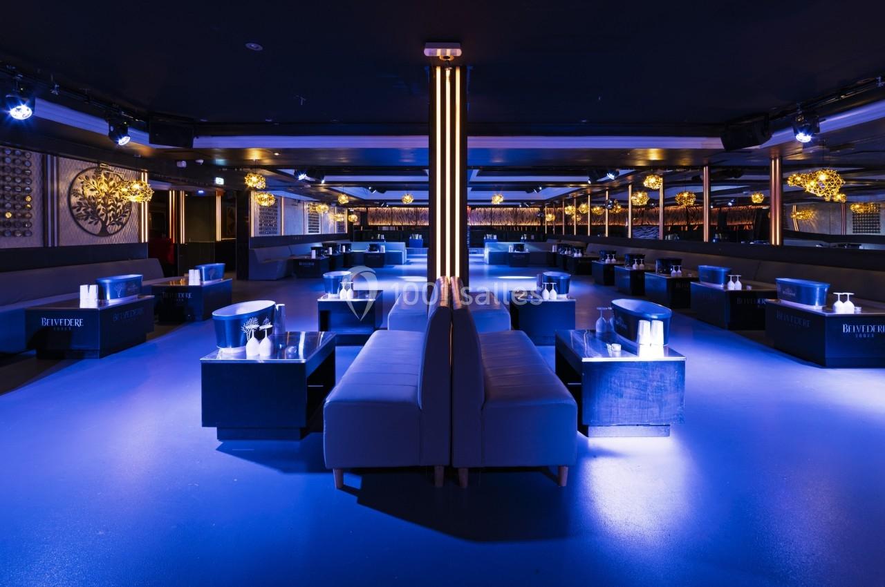 Salle de club avec éclairage tamisé bleu, banquettes, tables basses et décorations murales élégantes.