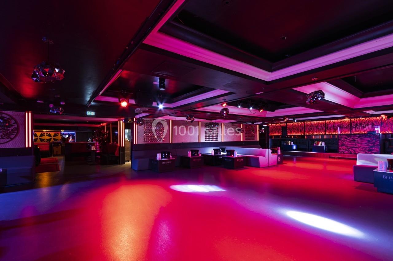 Salle de discothèque vide avec éclairage coloré, canapés et tables disposés autour d'une piste de danse.