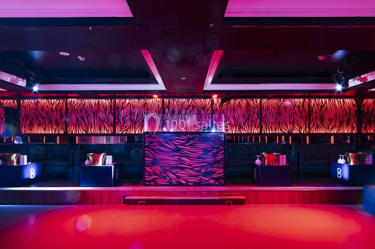 Salle de club avec éclairage rouge et violet, bar central décoré de motifs zébrés et banquettes en arrière-plan.