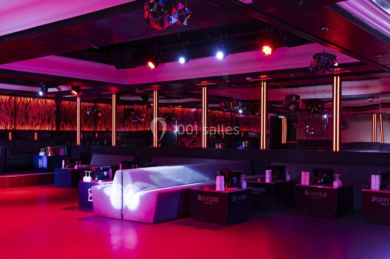 Salle de club avec éclairage rouge et violet, canapés blancs, tables basses et décorations modernes.