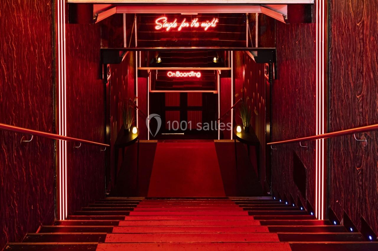 Escalier descendant dans un couloir éclairé par des néons rouges, avec des inscriptions lumineuses au fond.