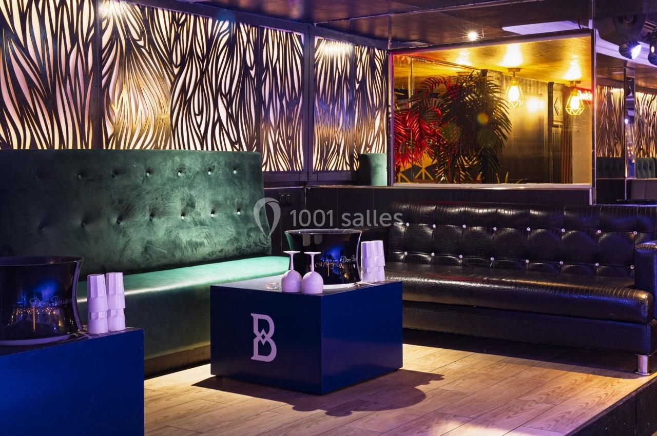 Salon avec canapés en cuir noir et vert, table basse bleue, éclairage tamisé et décor mural à motifs organiques.