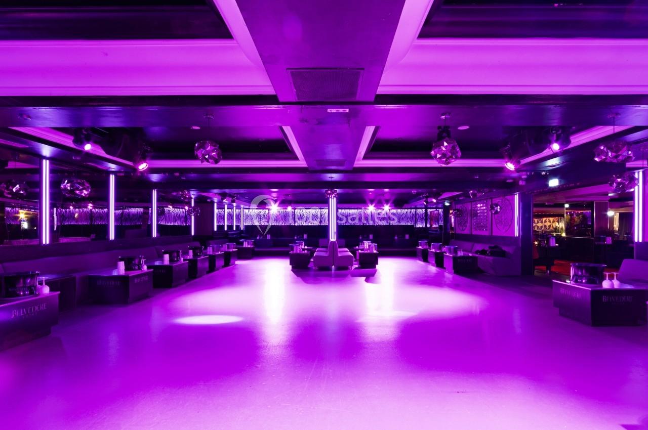 Salle de danse spacieuse éclairée par des lumières violettes, avec tables et banquettes disposées sur les côtés.