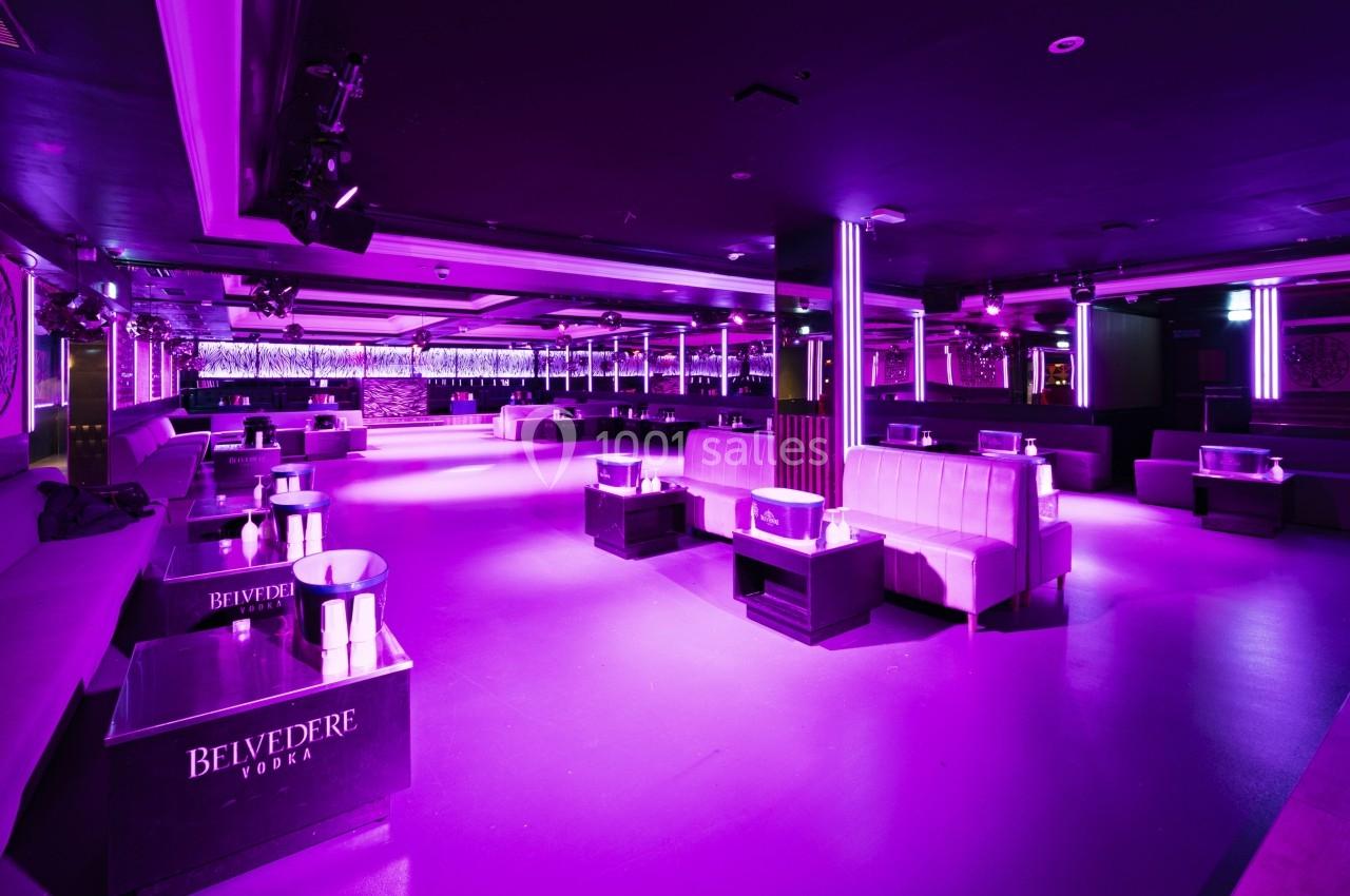 Salle de club avec éclairage violet, canapés blancs, tables basses et bar en arrière-plan.