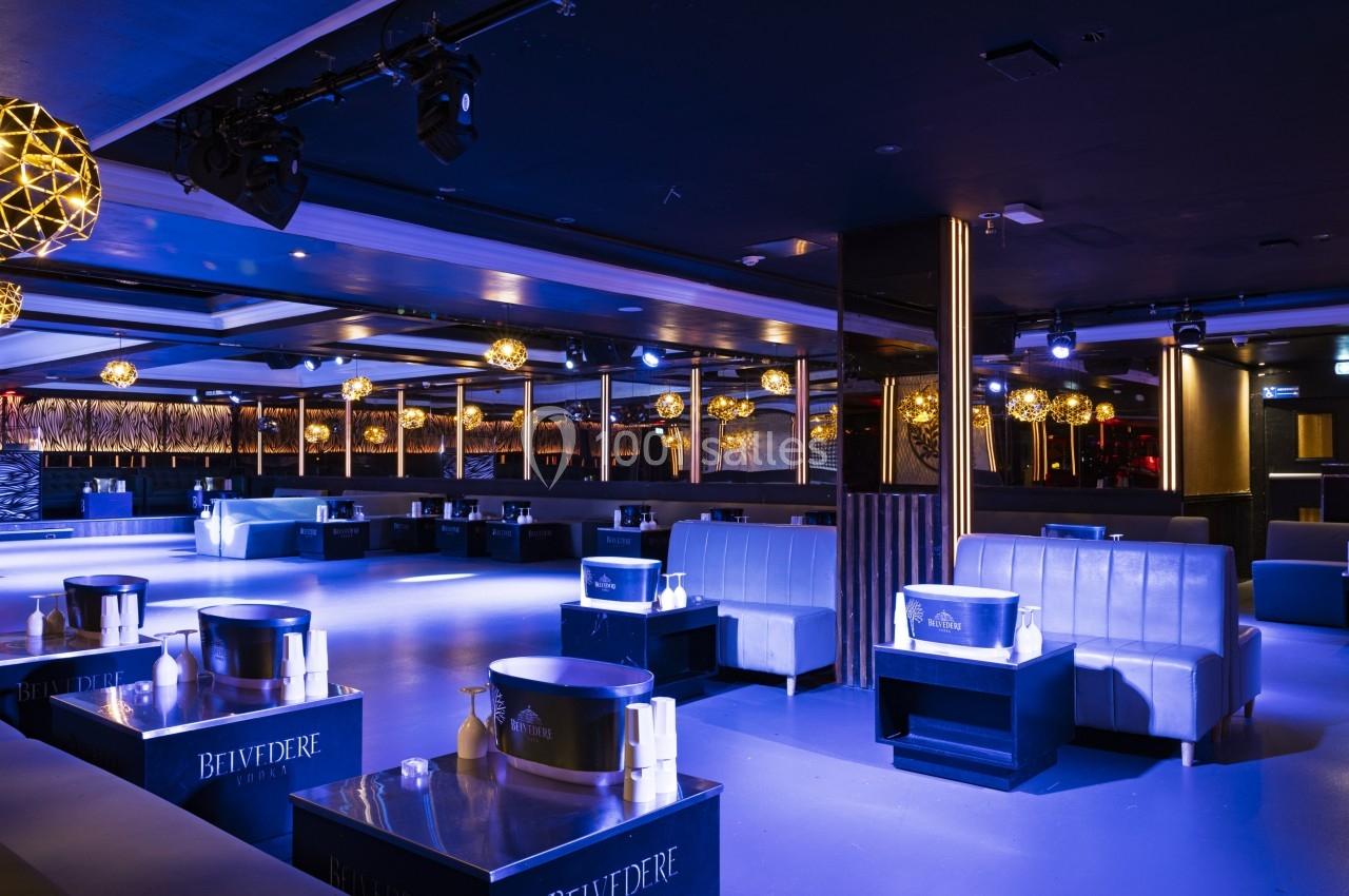 Salle de club moderne avec banquettes, tables basses et éclairage tamisé aux tons bleus et dorés.