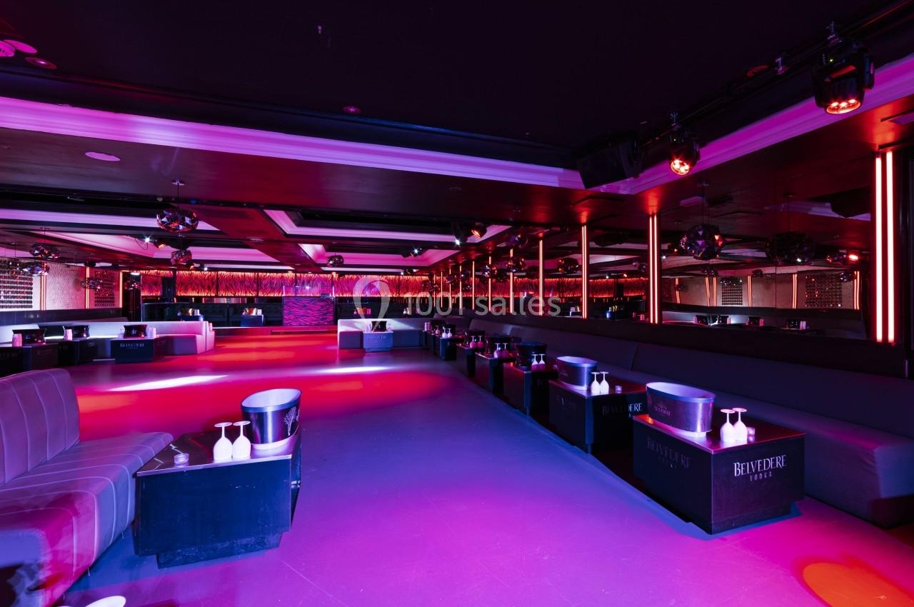 Salle de club vide avec éclairage coloré, banquettes, tables basses et murs ornés de miroirs.