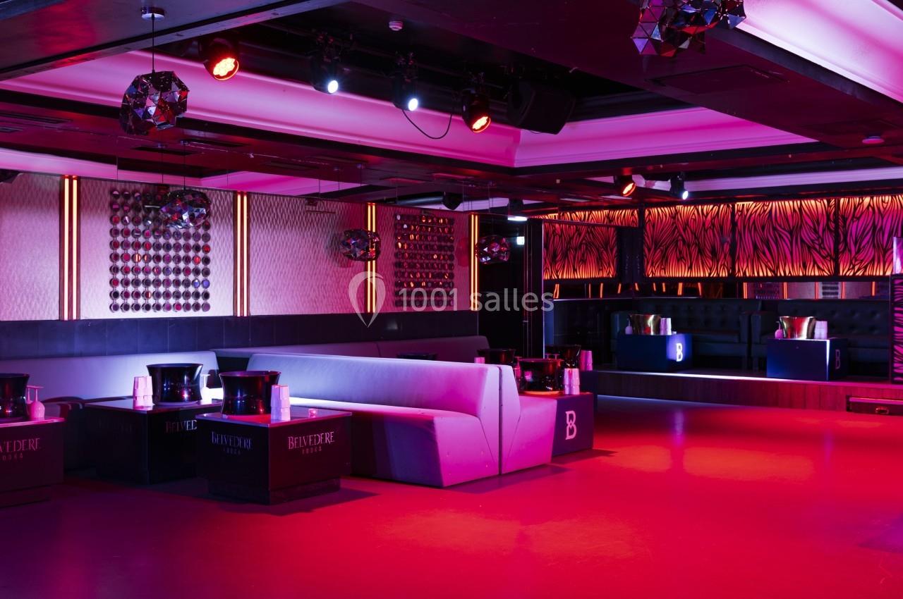 Salle de club avec éclairage rouge, banquettes grises, tables basses et décoration murale moderne.