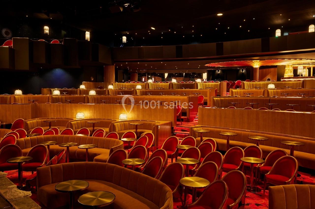 Salle de spectacle avec sièges rouges, tables rondes dorées et éclairage tamisé créant une ambiance chaleureuse.