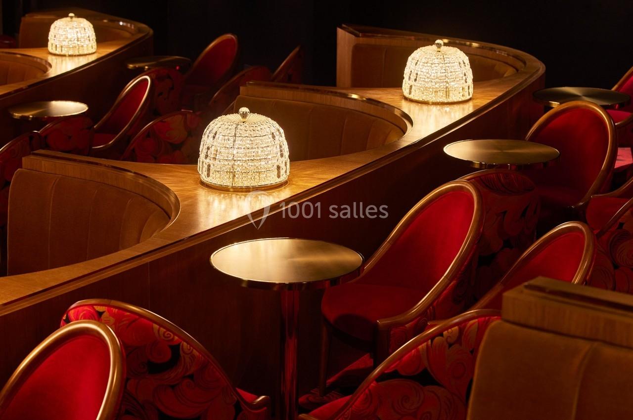Banquettes en velours rouge, tables rondes en bois et lampes élégantes dans une salle au décor chaleureux.