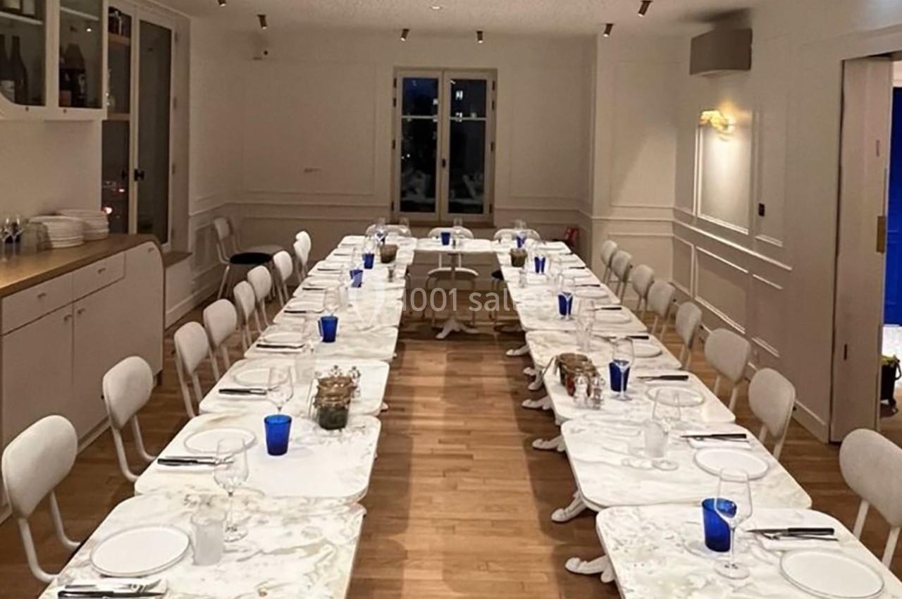 Salle à manger lumineuse avec deux longues tables blanches, chaises alignées, verres bleus et vaisselle disposée.