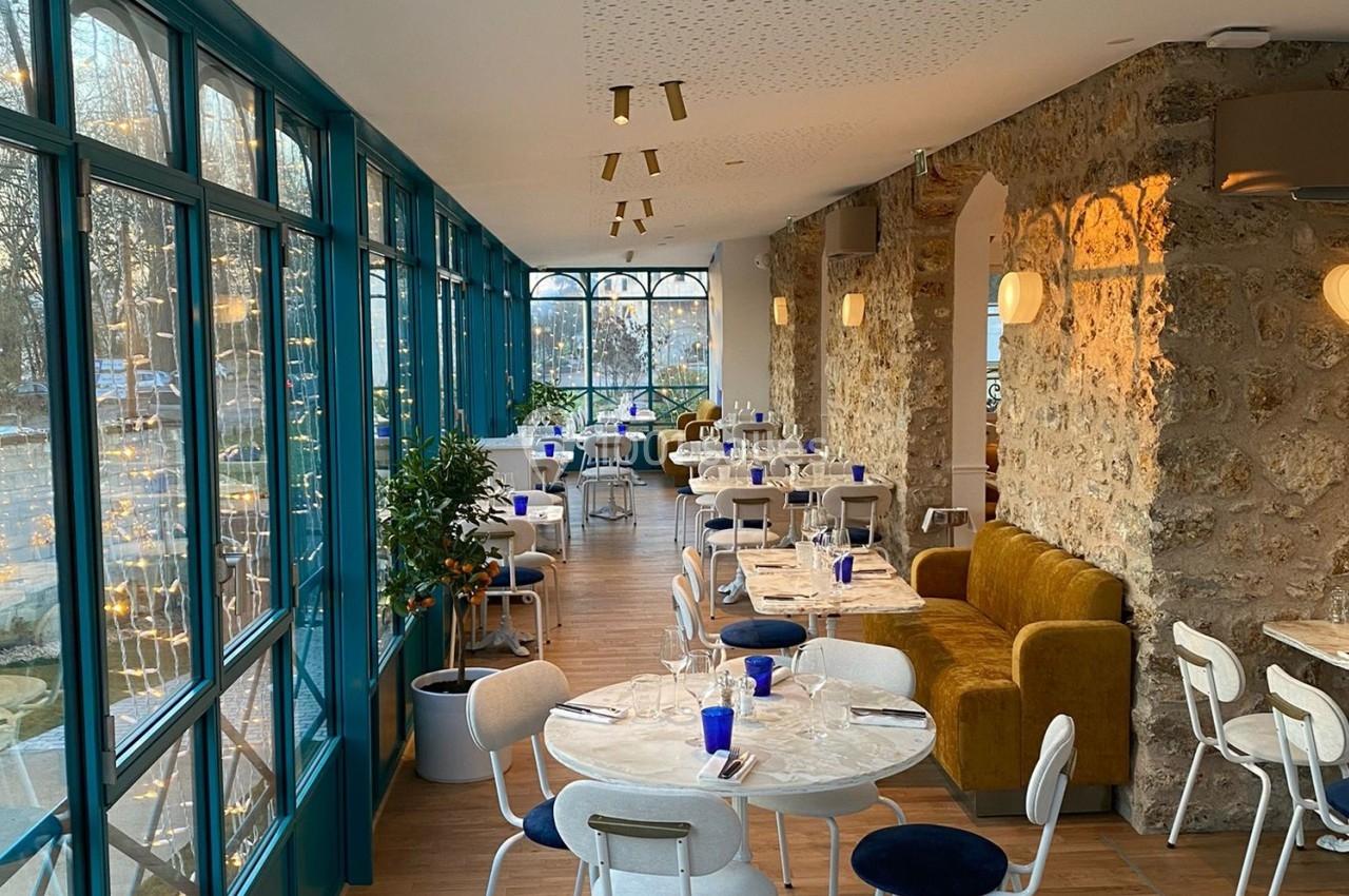 Salle de restaurant lumineuse avec murs en pierre, grandes baies vitrées, tables dressées et chaises blanches.