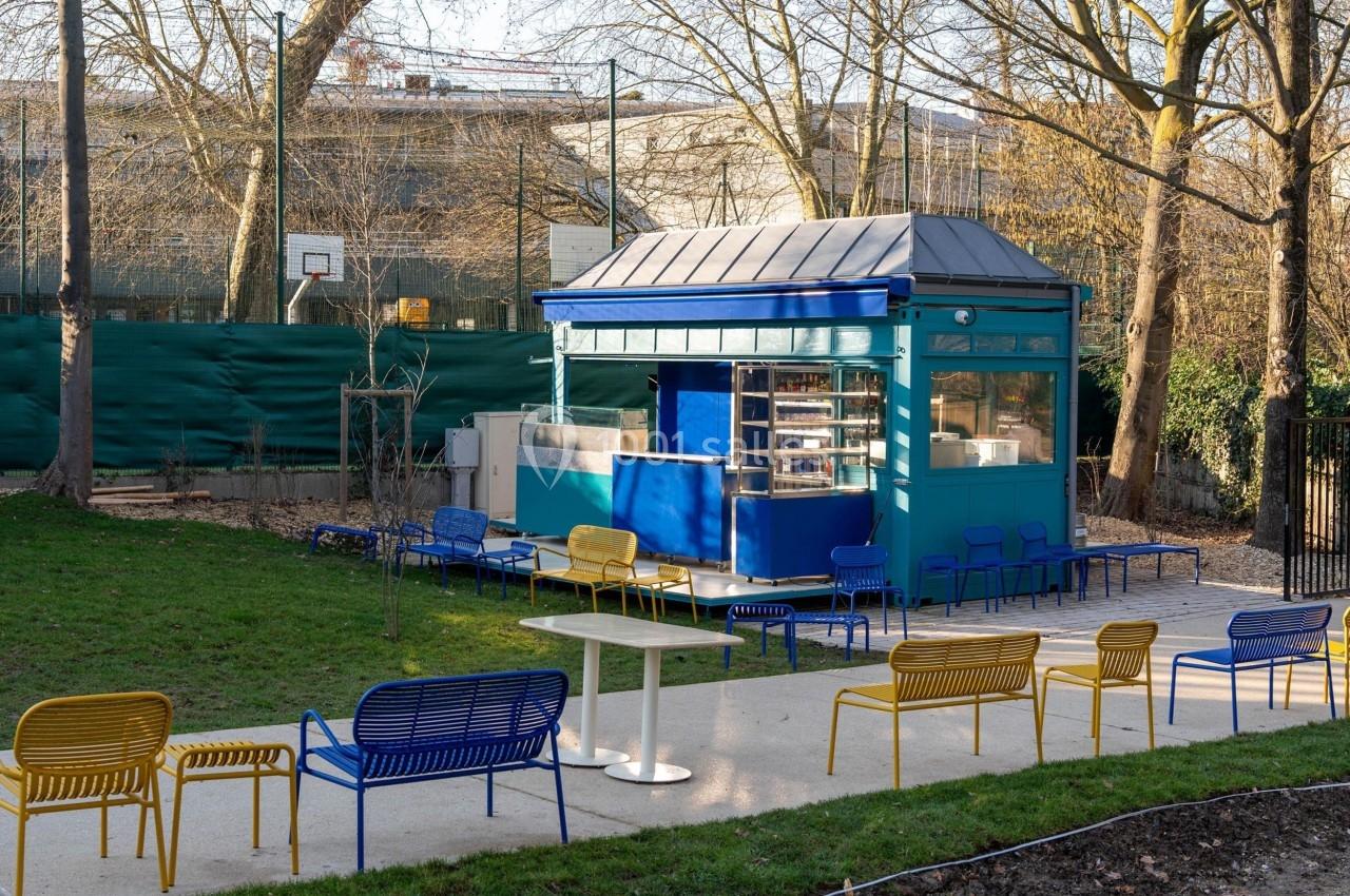 Kiosque bleu entouré de bancs colorés et de tables sur une allée, situé dans un parc avec arbres et terrain de sport.