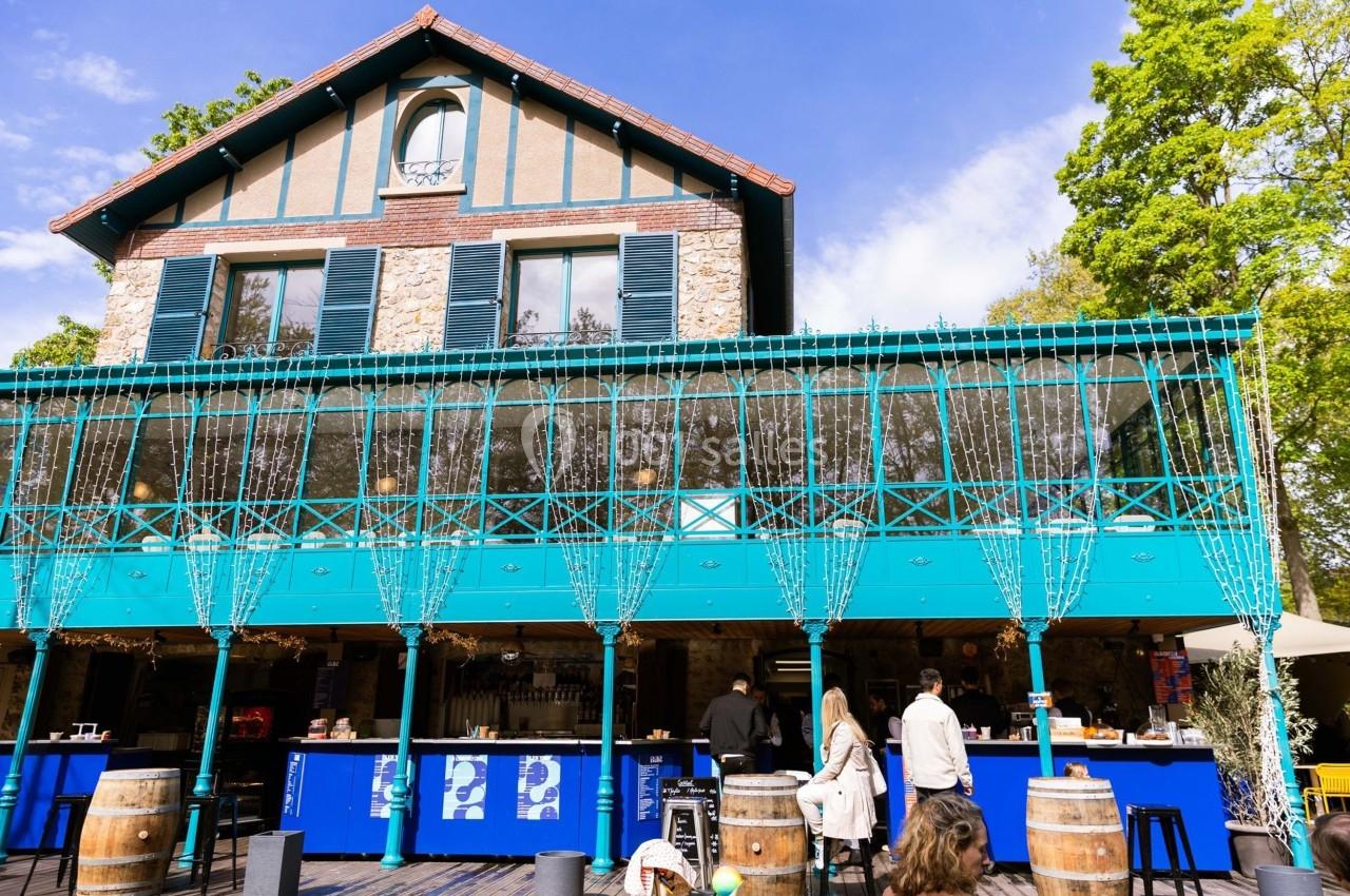 Façade d'un bâtiment en pierre avec terrasse vitrée et bar extérieur bleu, entouré de verdure.