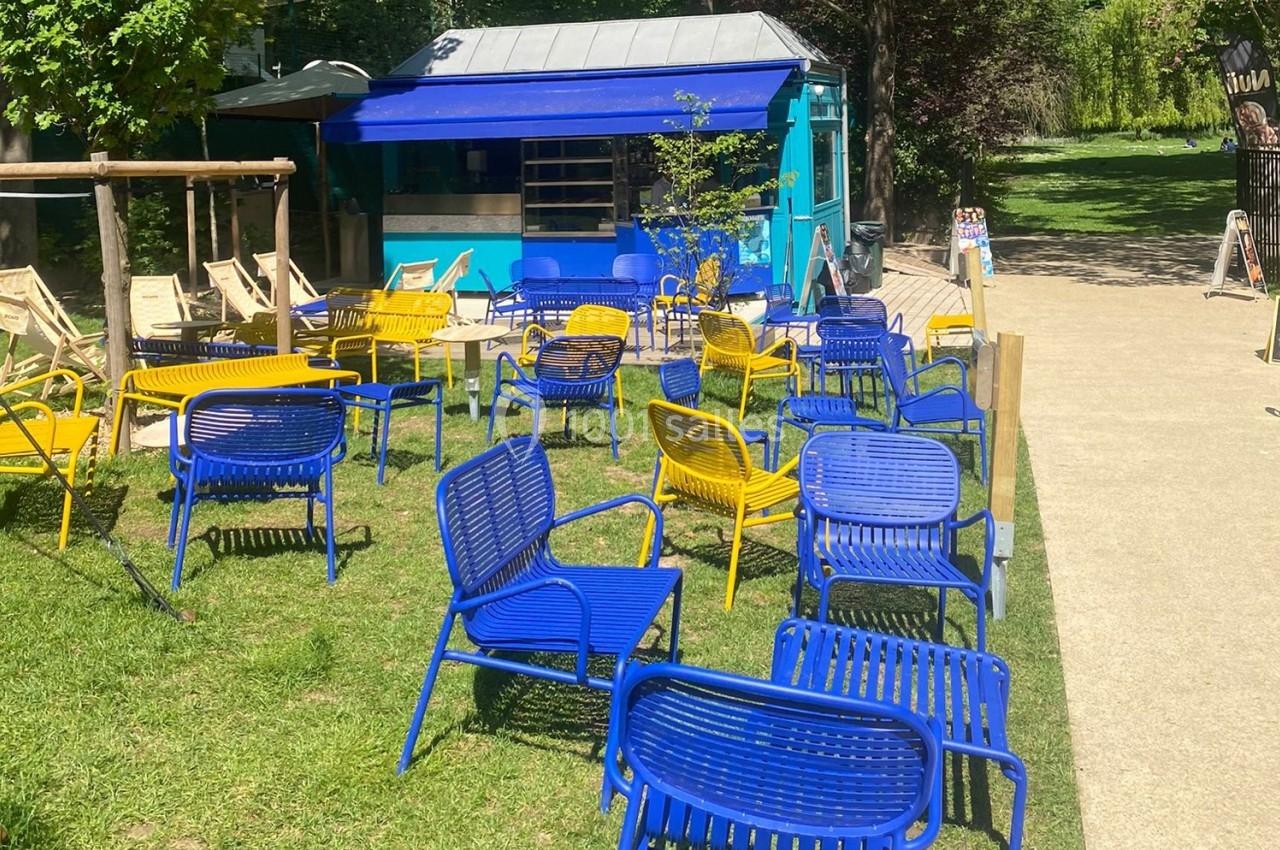 Chaises bleues et jaunes disposées sur une pelouse devant un kiosque bleu, entouré d'arbres et d'un chemin pavé.