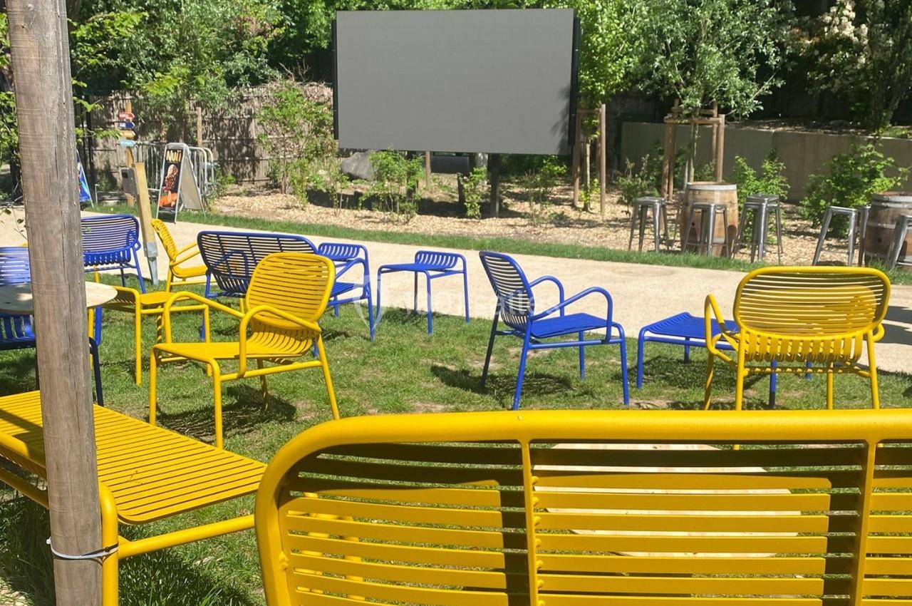 Chaises et bancs colorés disposés en plein air devant un écran, entourés de verdure et d'arbres.