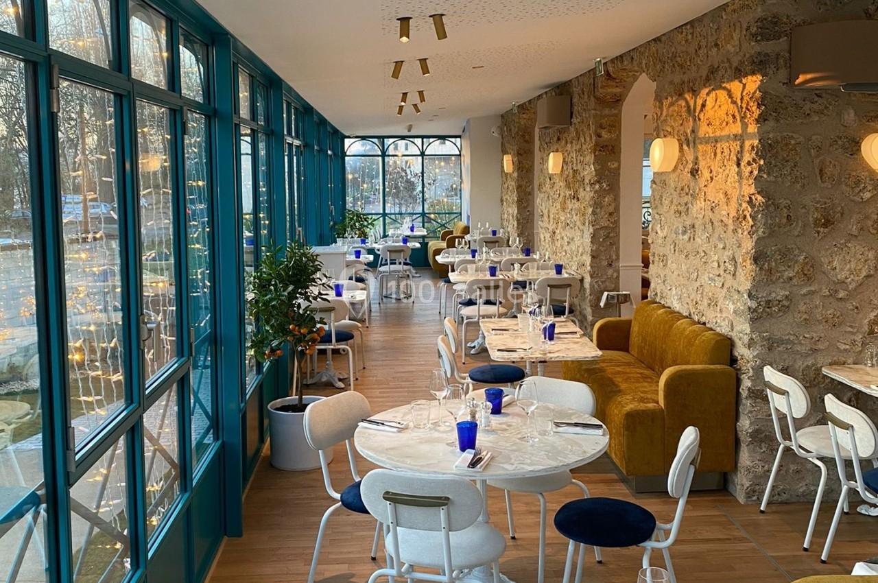 Salle de restaurant lumineuse avec murs en pierre, grandes baies vitrées, tables rondes et chaises blanches.