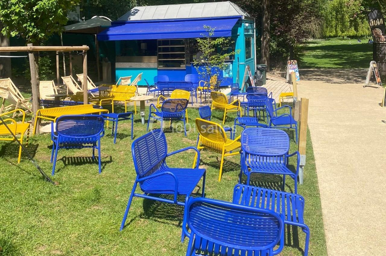 Chaises bleues et jaunes disposées sur une pelouse devant un kiosque bleu dans un parc ensoleillé.