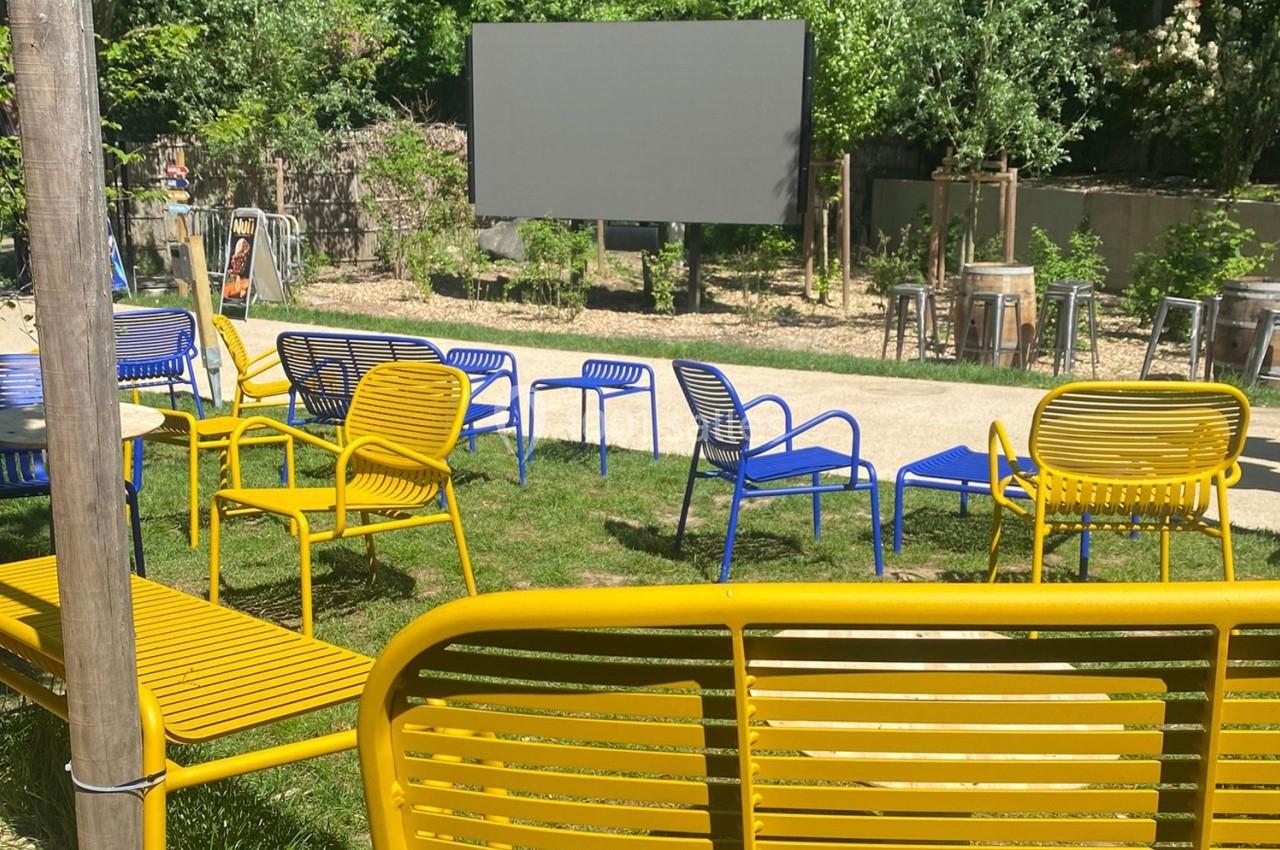Chaises et bancs colorés disposés en extérieur devant un écran, entourés de verdure et de lumière naturelle.