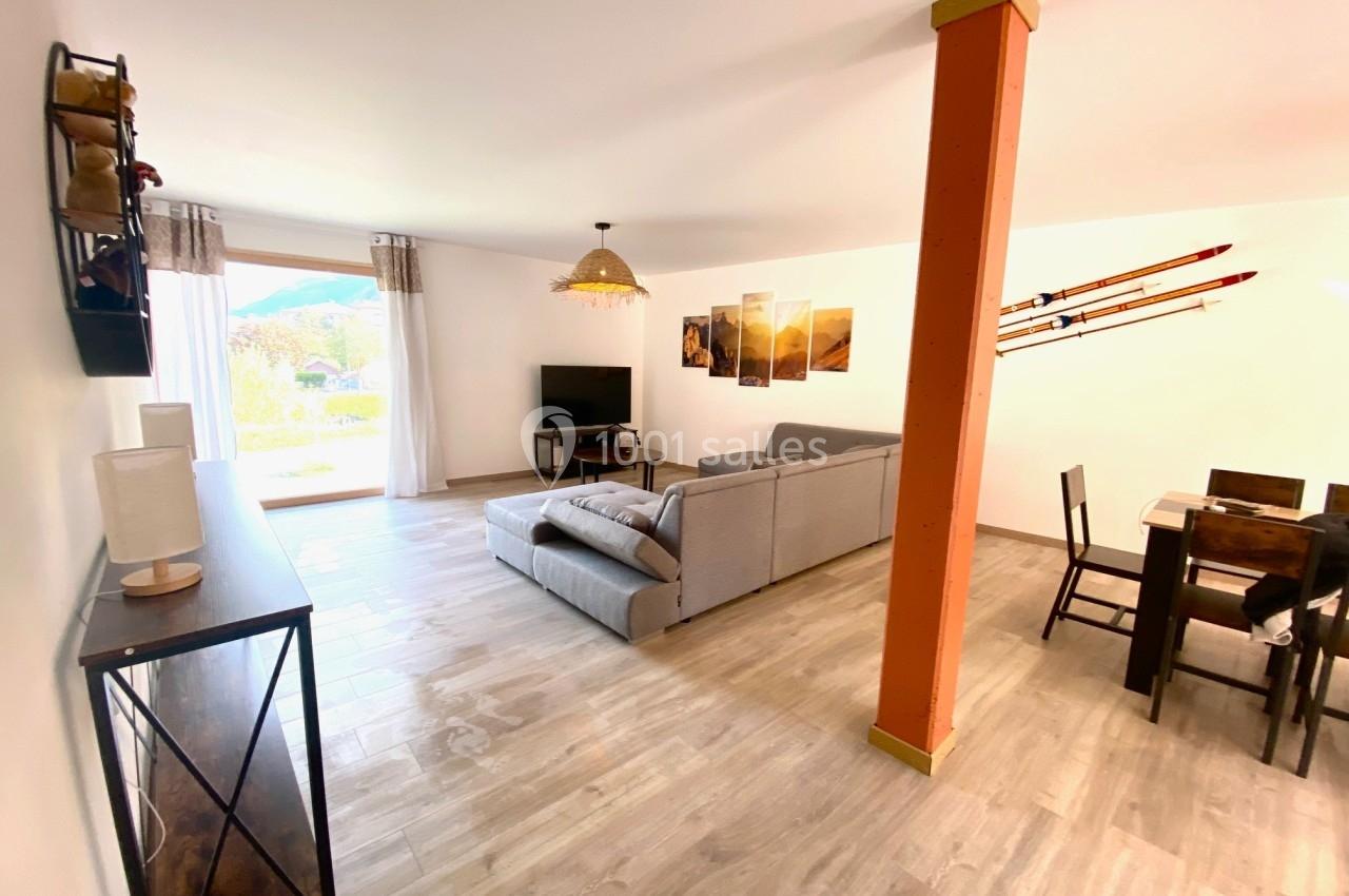 Salon lumineux avec canapé gris, table à manger en bois, skis décoratifs au mur et grande baie vitrée donnant sur l…