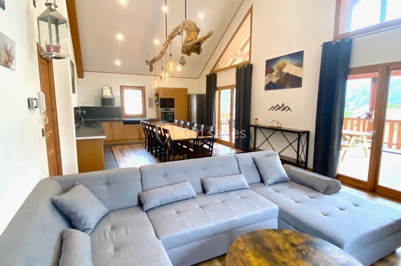 Salon spacieux avec canapé d'angle gris, table basse ronde en bois, cuisine ouverte et grande baie vitrée.