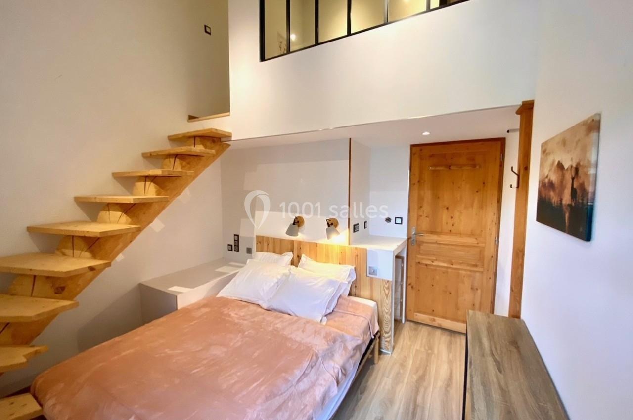 Chambre lumineuse avec lit double, escalier en bois menant à une mezzanine, et décoration minimaliste.