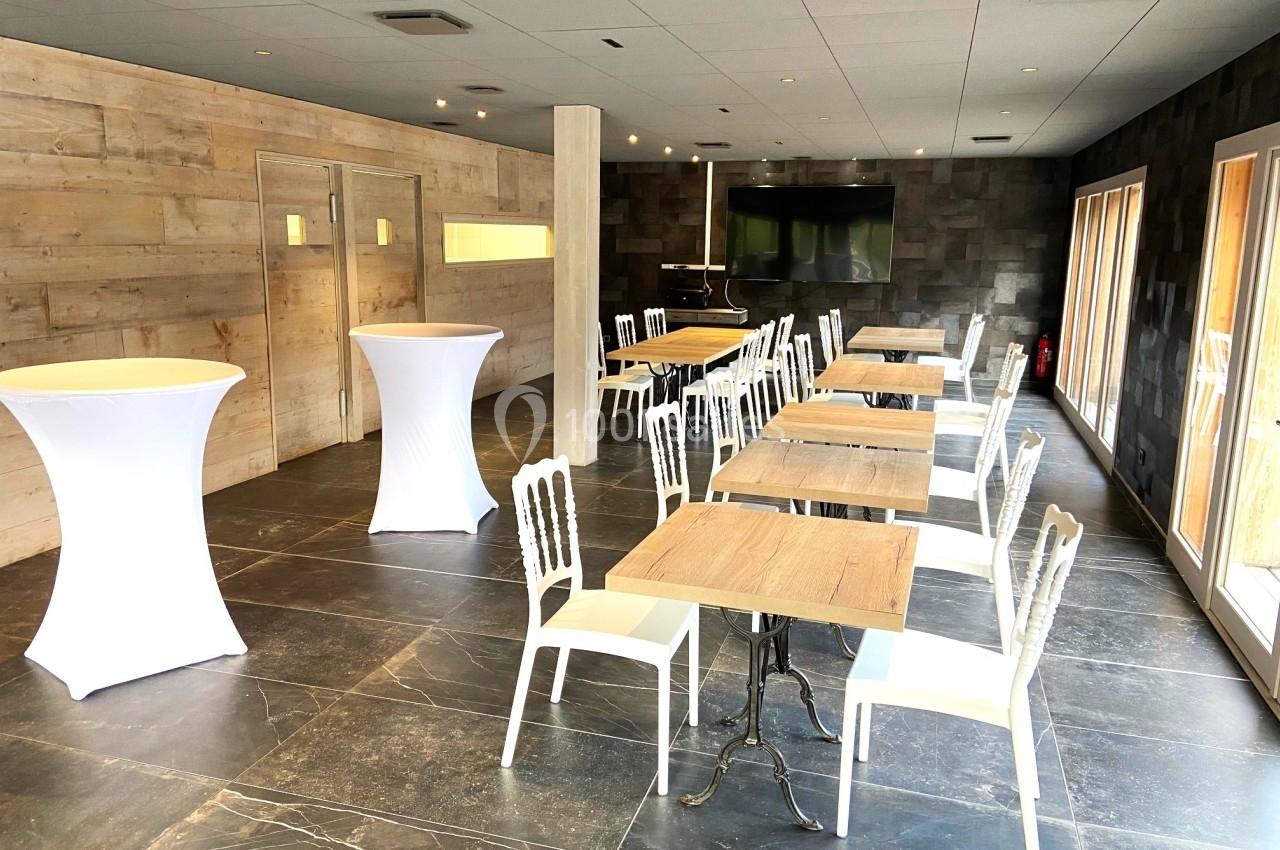 Salle lumineuse avec tables en bois, chaises blanches, mange-debout et grandes fenêtres laissant entrer la lumière naturelle.