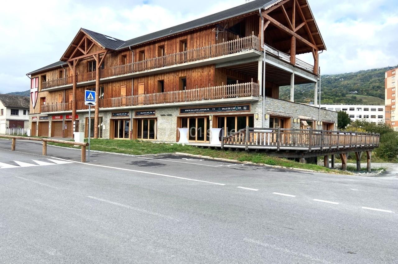 Bâtiment en bois et pierre à deux étages avec balcons, situé près d'une route et entouré de montagnes.