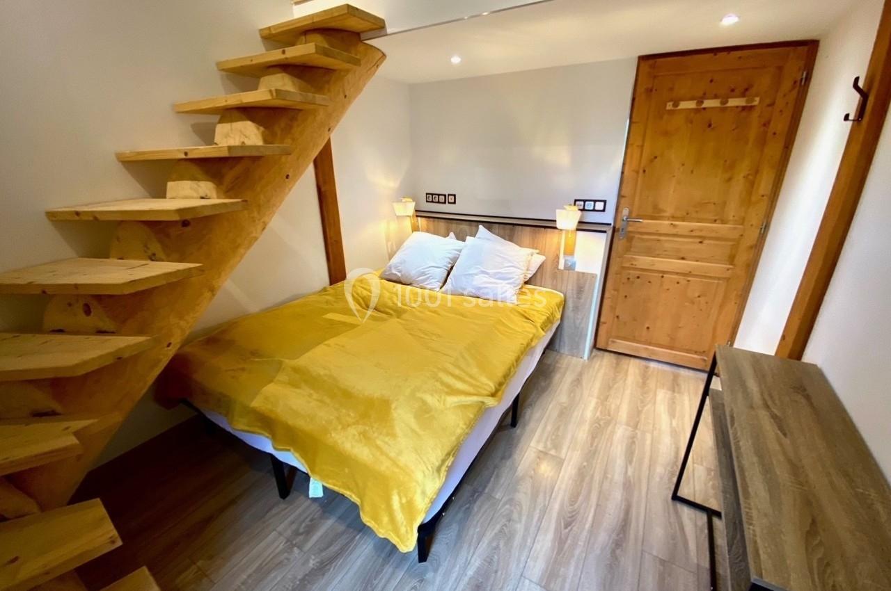 Chambre avec lit double, escalier en bois menant à une mezzanine, sol en parquet et éclairage chaleureux.