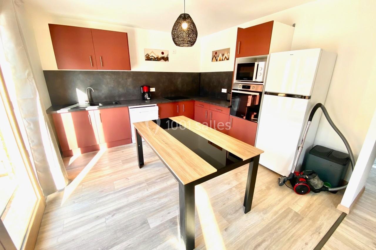 Cuisine moderne avec meubles rouges, table en bois, électroménagers intégrés, réfrigérateur blanc et aspirateur au sol.