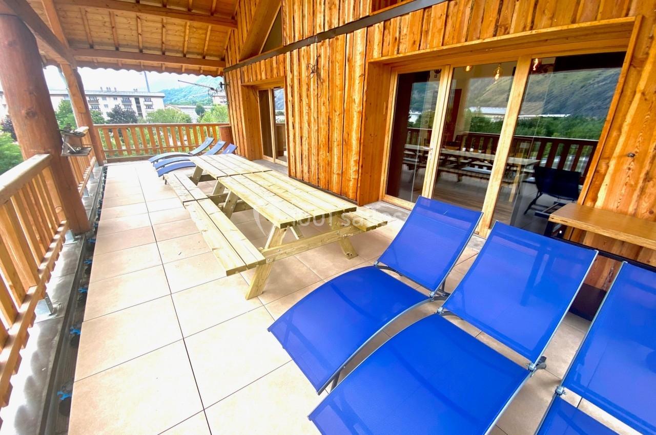 Terrasse en bois avec table de pique-nique et chaises longues bleues, offrant une vue sur des montagnes et des bâtiments.