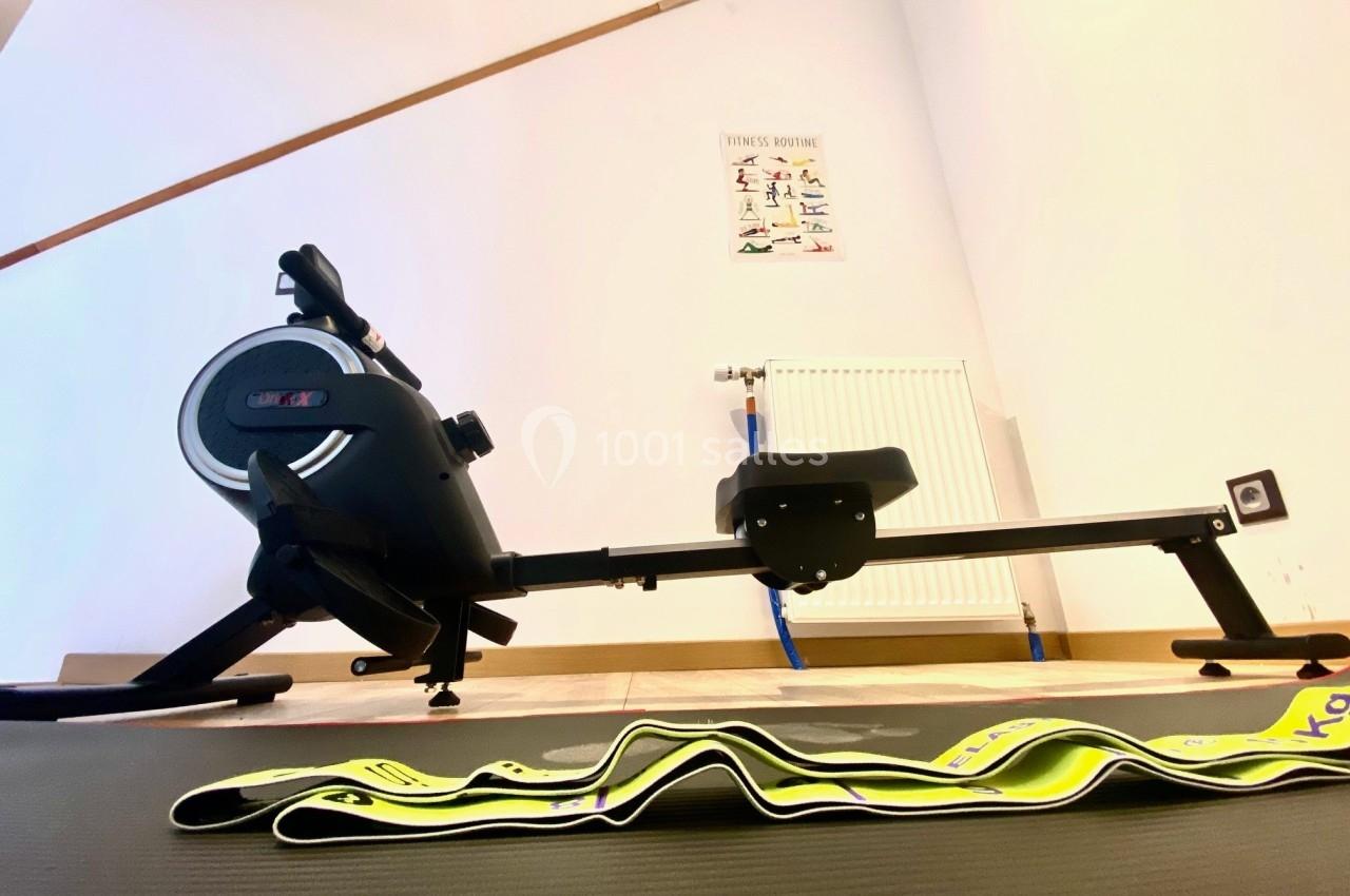 Rameur d'intérieur noir placé dans une pièce lumineuse avec un tapis de sol et des bandes élastiques au premier plan.