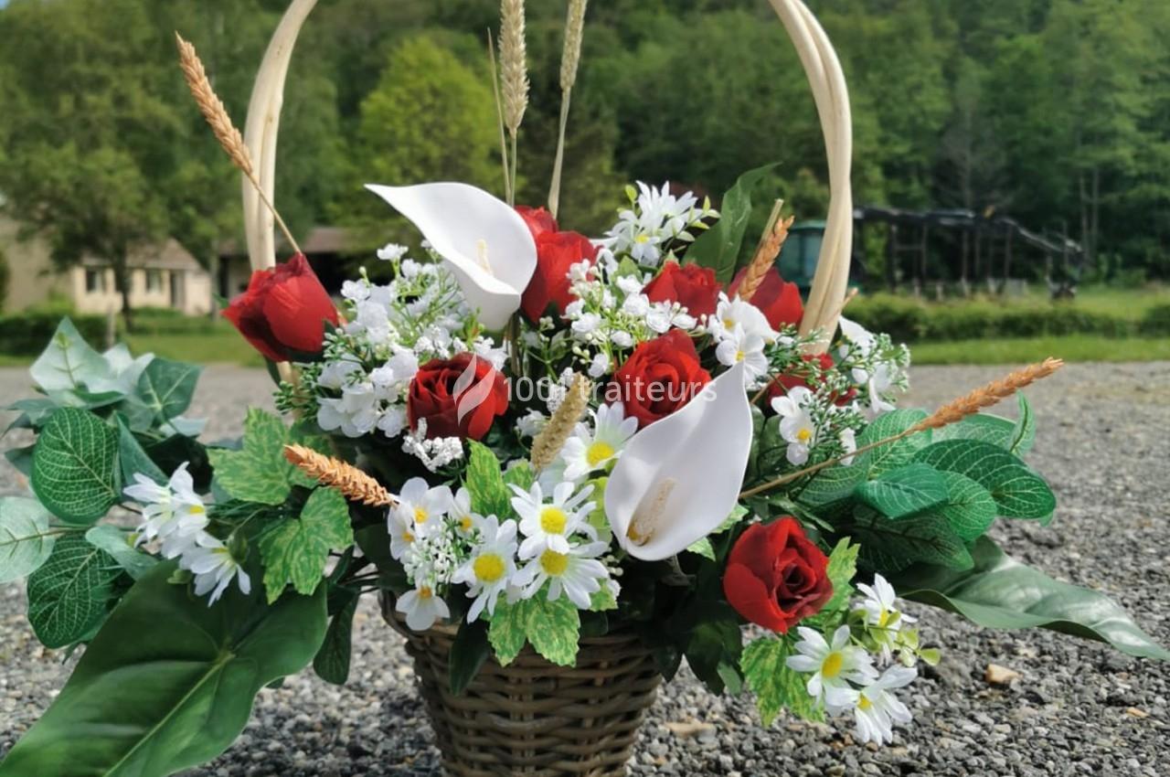 Panier en osier rempli de roses rouges, arums blancs, marguerites et feuillage, posé sur un sol gravillonné en extérieur.