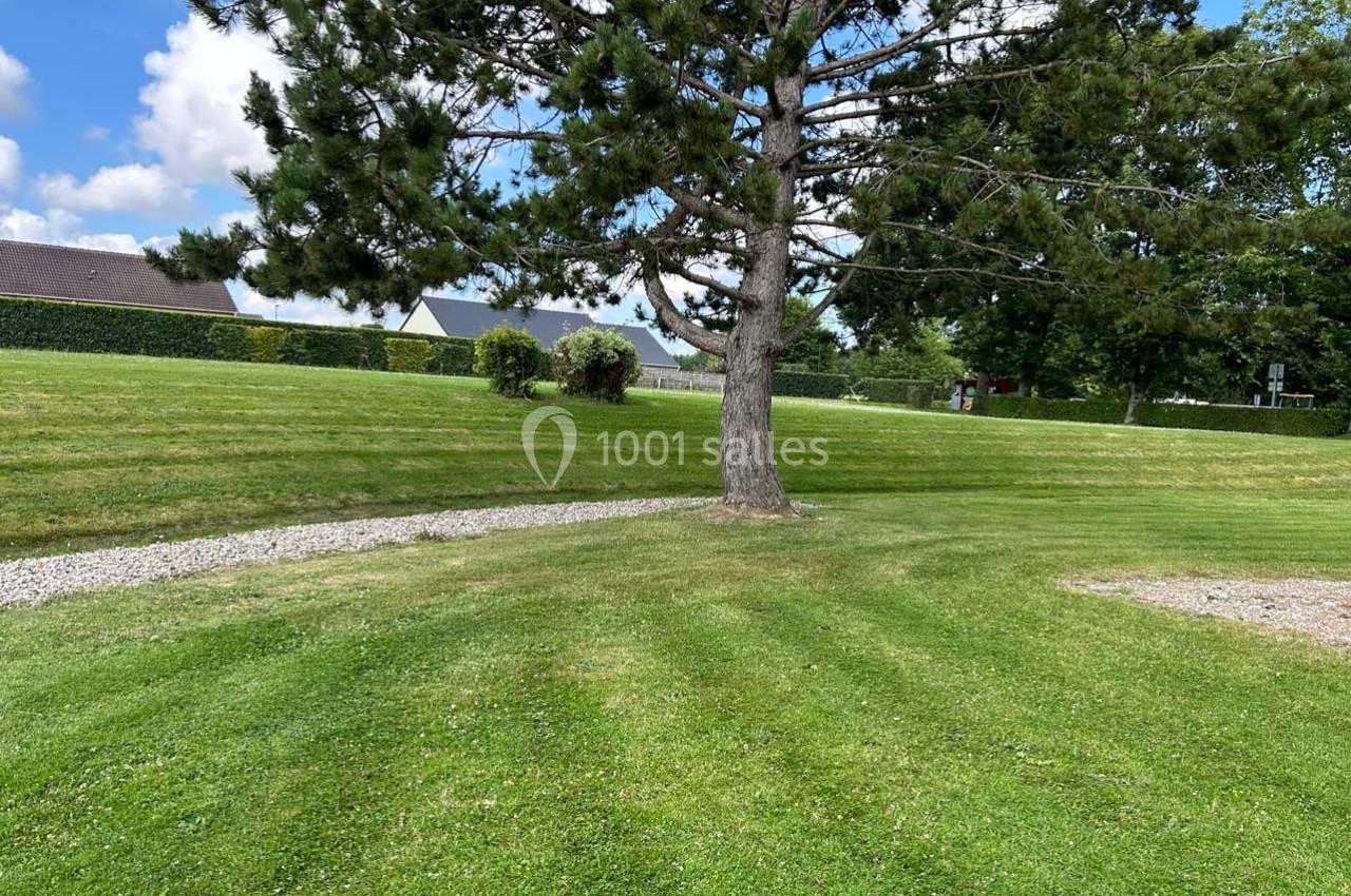 Pelouse verte avec un grand arbre au centre, chemin de gravier et haies en arrière-plan sous un ciel partiellement nuageux.