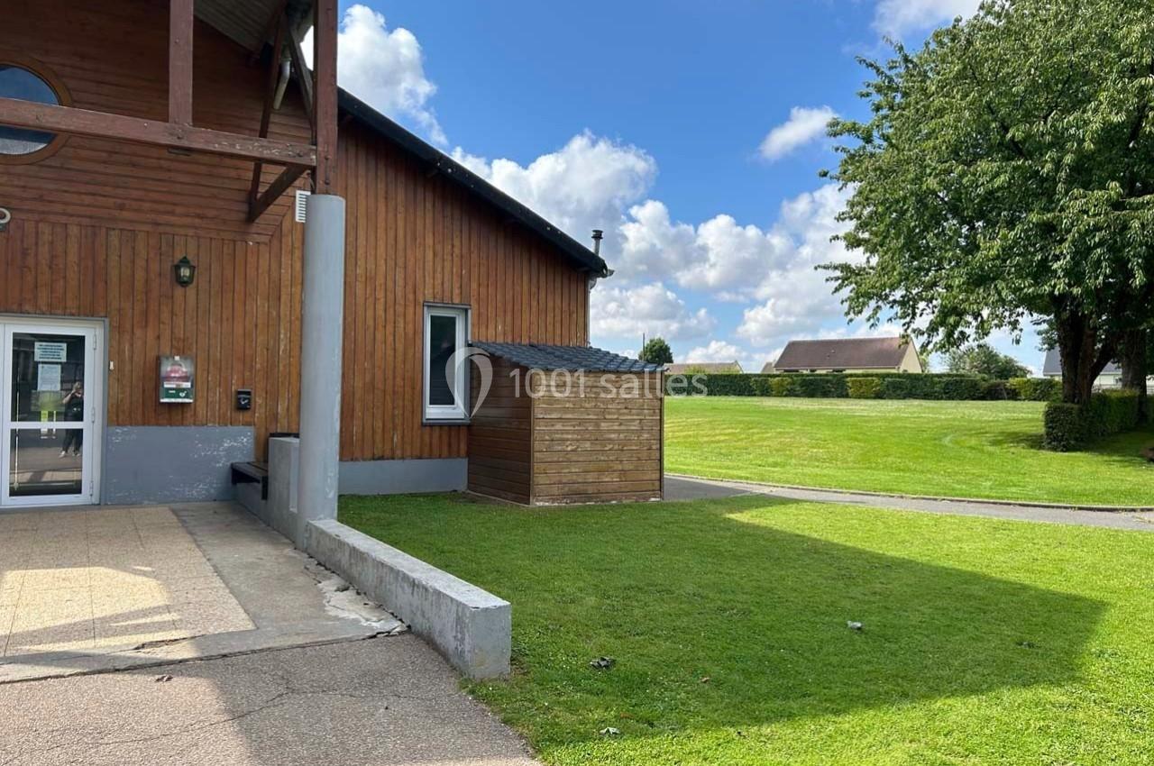 Bâtiment en bois avec une entrée vitrée, pelouse et arbres sous un ciel partiellement nuageux.