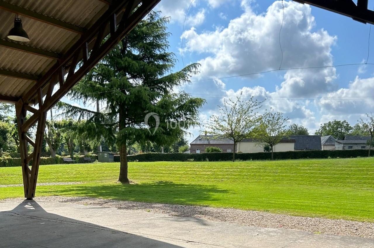 Vue d'un espace extérieur avec pelouse, arbres et bâtiments en arrière-plan, prise depuis un abri couvert.