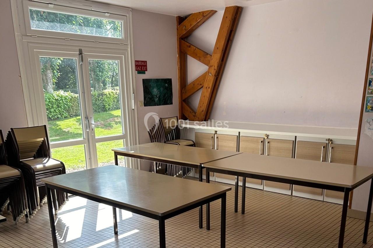Salle lumineuse avec tables et chaises empilées, carrelage au sol, grande porte vitrée donnant sur un espace vert.