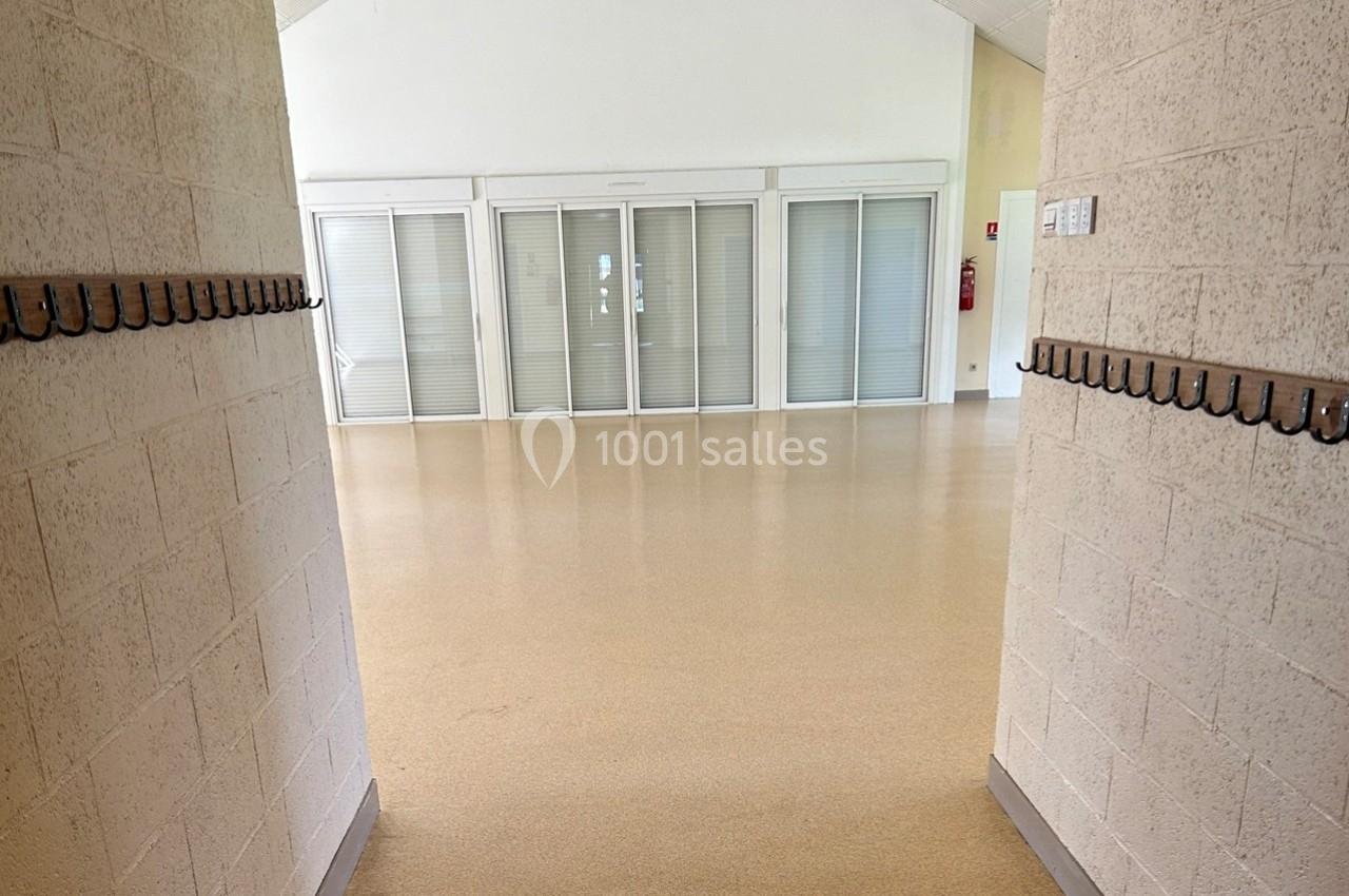 Vue d'une salle vide avec sol beige, murs clairs, crochets muraux et baies vitrées fermées par des stores.