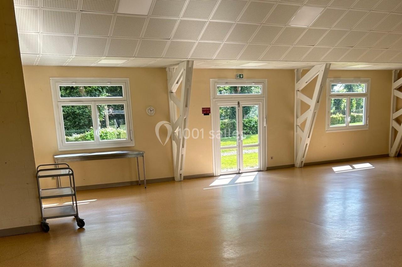 Salle lumineuse avec sol beige, murs clairs, fenêtres donnant sur un espace vert, table et chariot en métal à gauche.