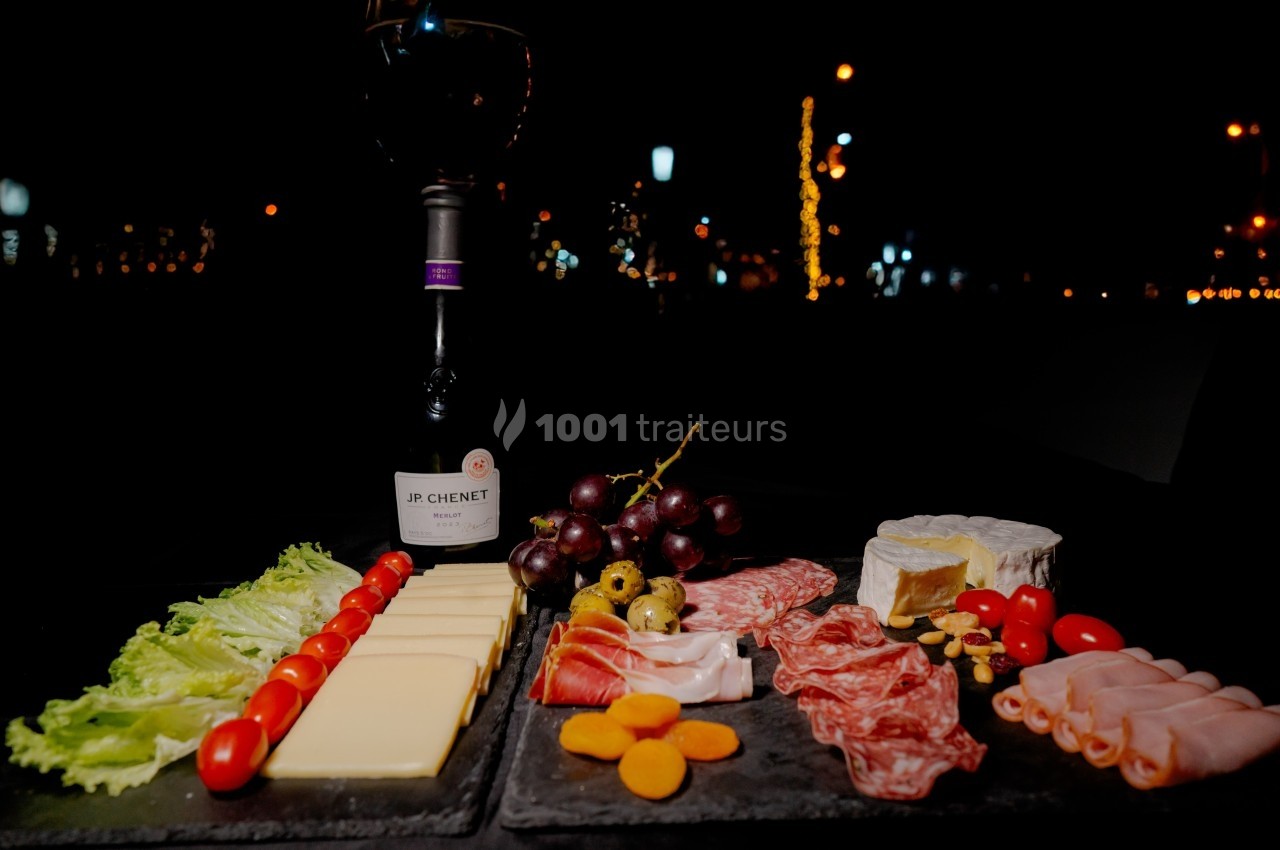 Plateau de charcuterie et fromages avec fruits, légumes, et une bouteille de vin rouge, sur une table en extérieur la nuit.
