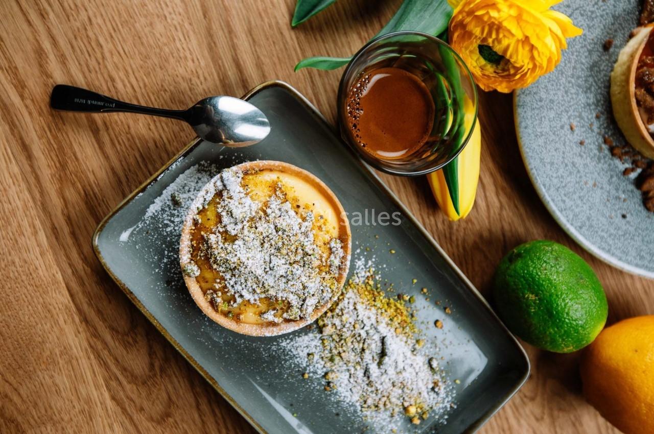 Tartelette saupoudrée de sucre glace sur une assiette, accompagnée d'un café, d'agrumes et de fleurs sur une table en bois.