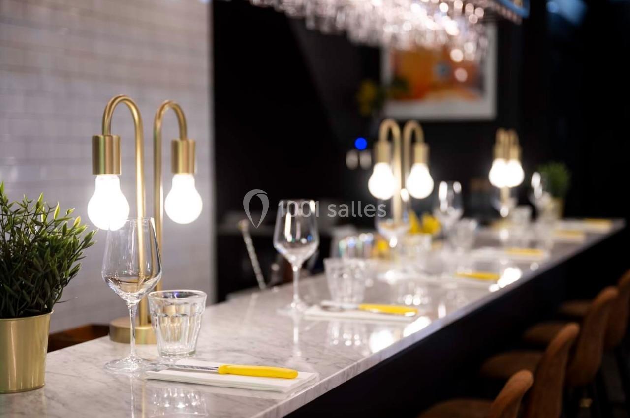 Comptoir en marbre avec couverts, verres et lampes décoratives, entouré de tabourets dans un espace lumineux.