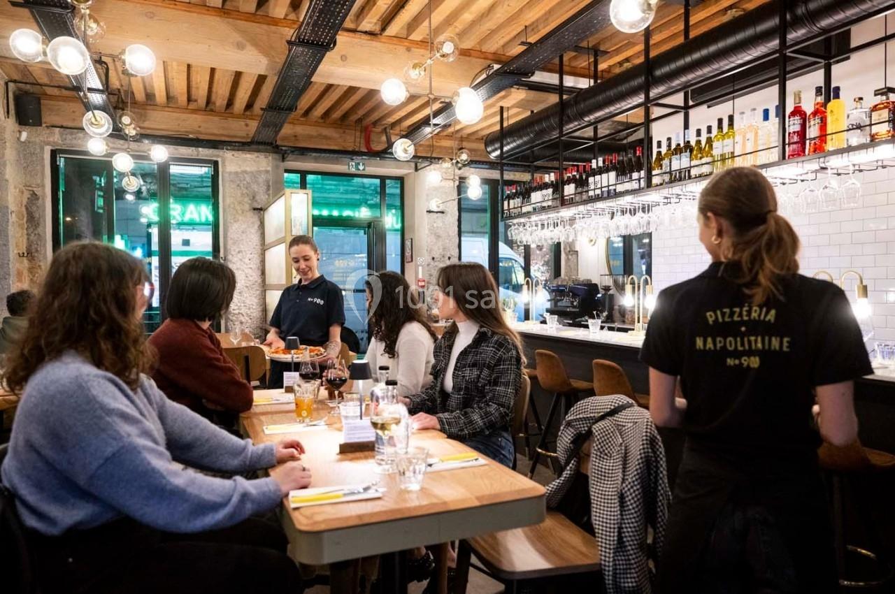 Serveuses interagissant avec des clients attablés dans une pizzeria au décor moderne et chaleureux.