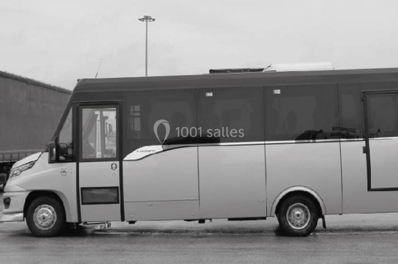 Minibus stationné sur une surface goudronnée, vu de côté, avec un bâtiment et un lampadaire en arrière-plan.