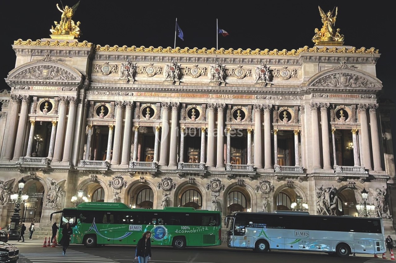Façade illuminée de l'Opéra Garnier à Paris la nuit, avec deux bus stationnés devant.