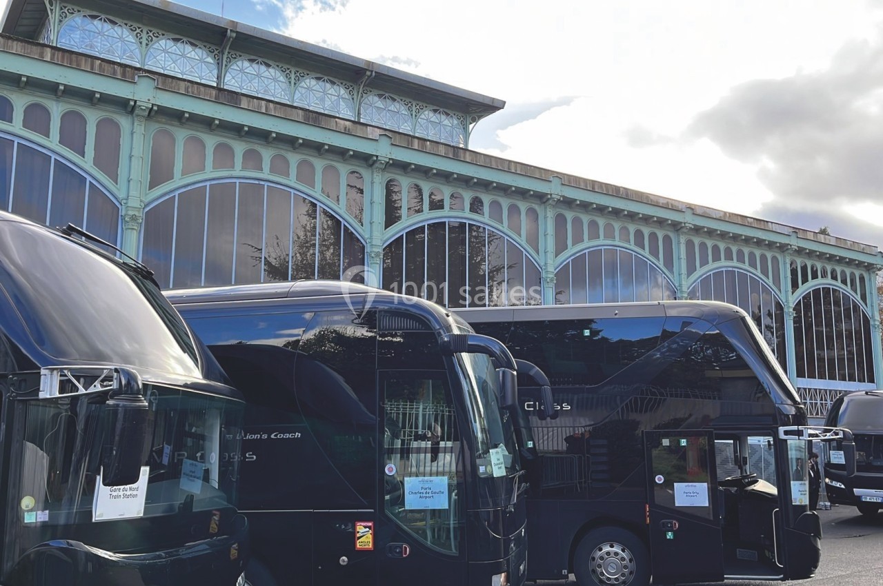 Autocars stationnés devant un bâtiment à structure métallique et verrière, sous un ciel partiellement nuageux.