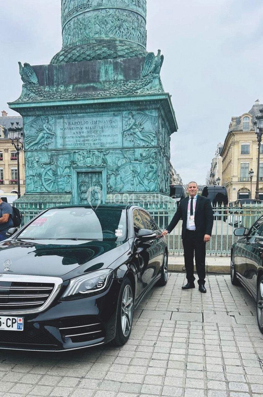 Un homme en costume se tient près d'une voiture noire stationnée devant la colonne Vendôme à Paris.