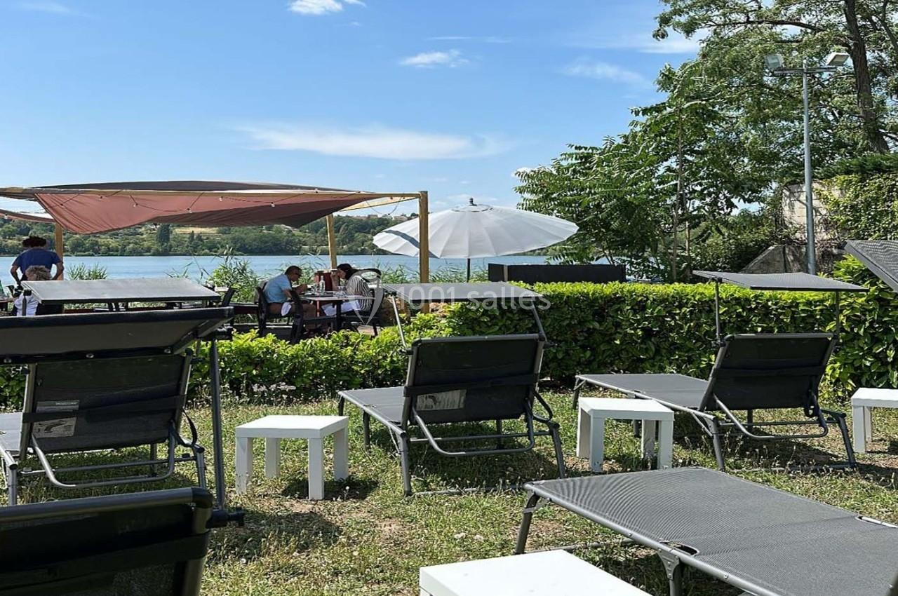 Chaises longues et tables disposées sur une pelouse près d'un lac, avec des parasols et une pergola en arrière-plan.