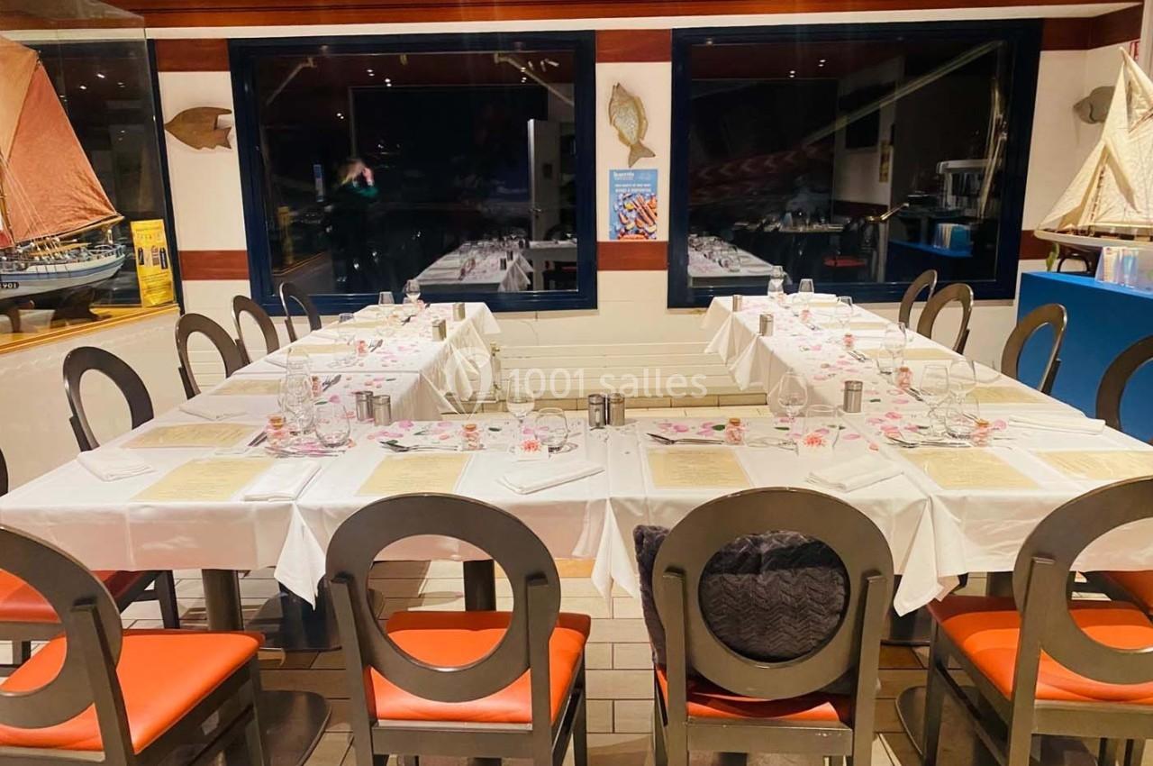 Salle de restaurant avec tables dressées, nappes blanches, chaises noires et décorations maritimes en arrière-plan.